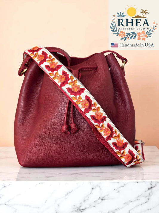 Red leather bucket bag with a floral purse strap on a marble surface, featuring the RHEA brand.