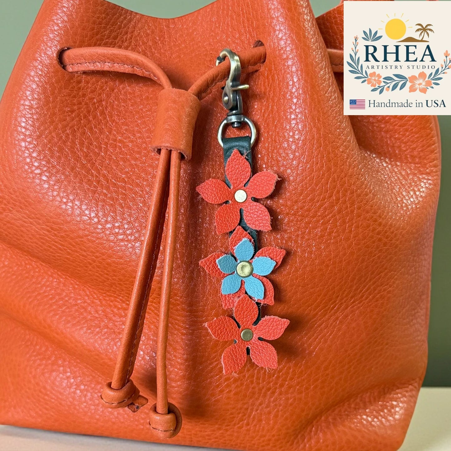 Orange leather bag with a floral keychain charm and RHEA Artistry Studio logo.