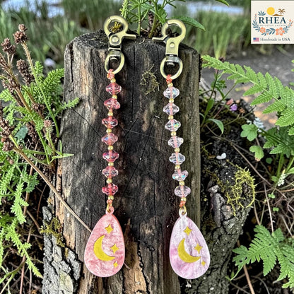 Pair of charms with pink teardrop designs and yellow crescent moons hanging on a wooden block with greenery in the background.