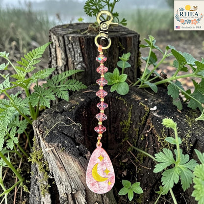 Decorative keychain with pink teardrop design hanging on a tree stump with plants around