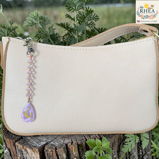 Beige handbag with a decorative keychain on a wooden surface with plants in the background, featuring the RHEA brand.