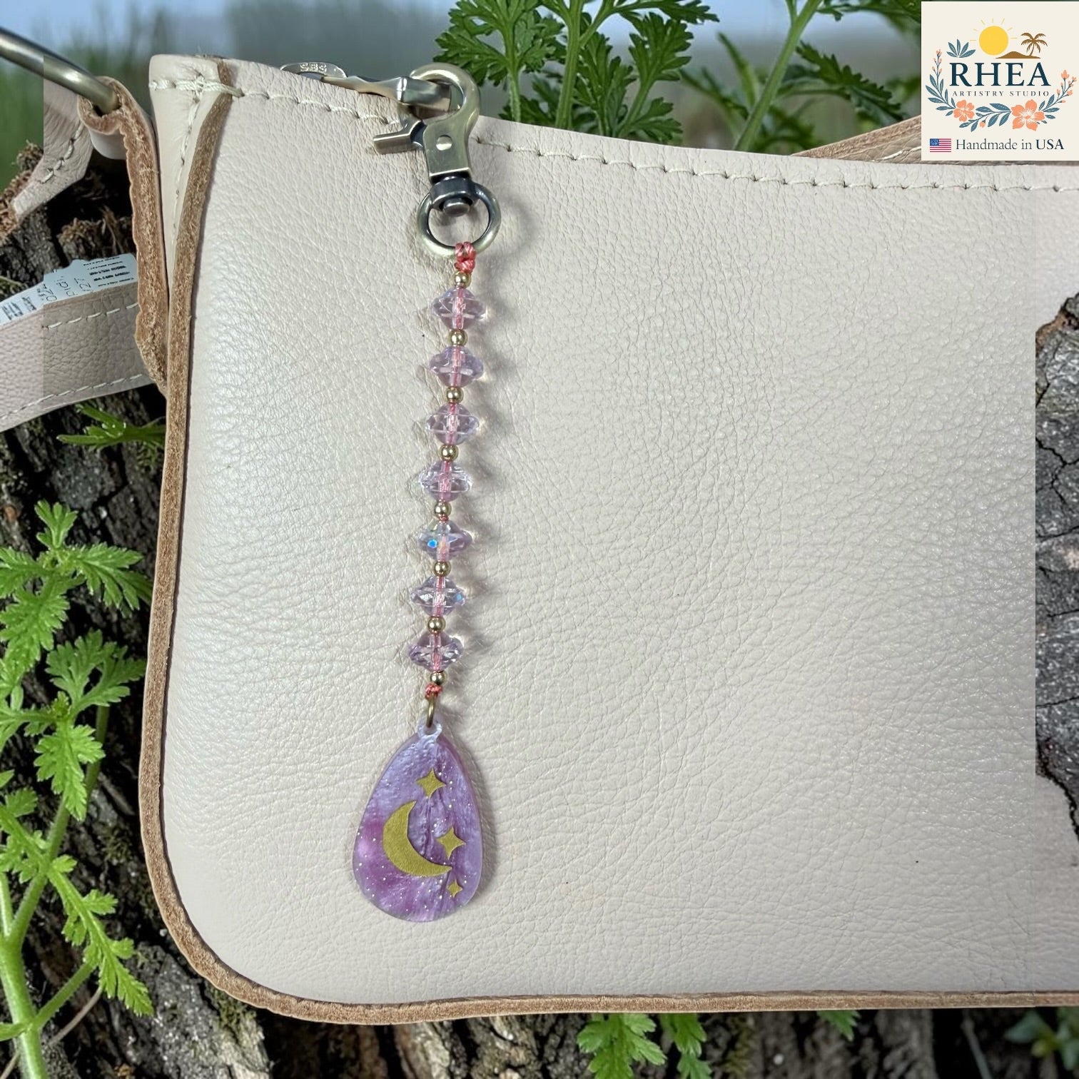 Decorative keychain with purple teardrop charm on a white leather bag with green plants in the background.