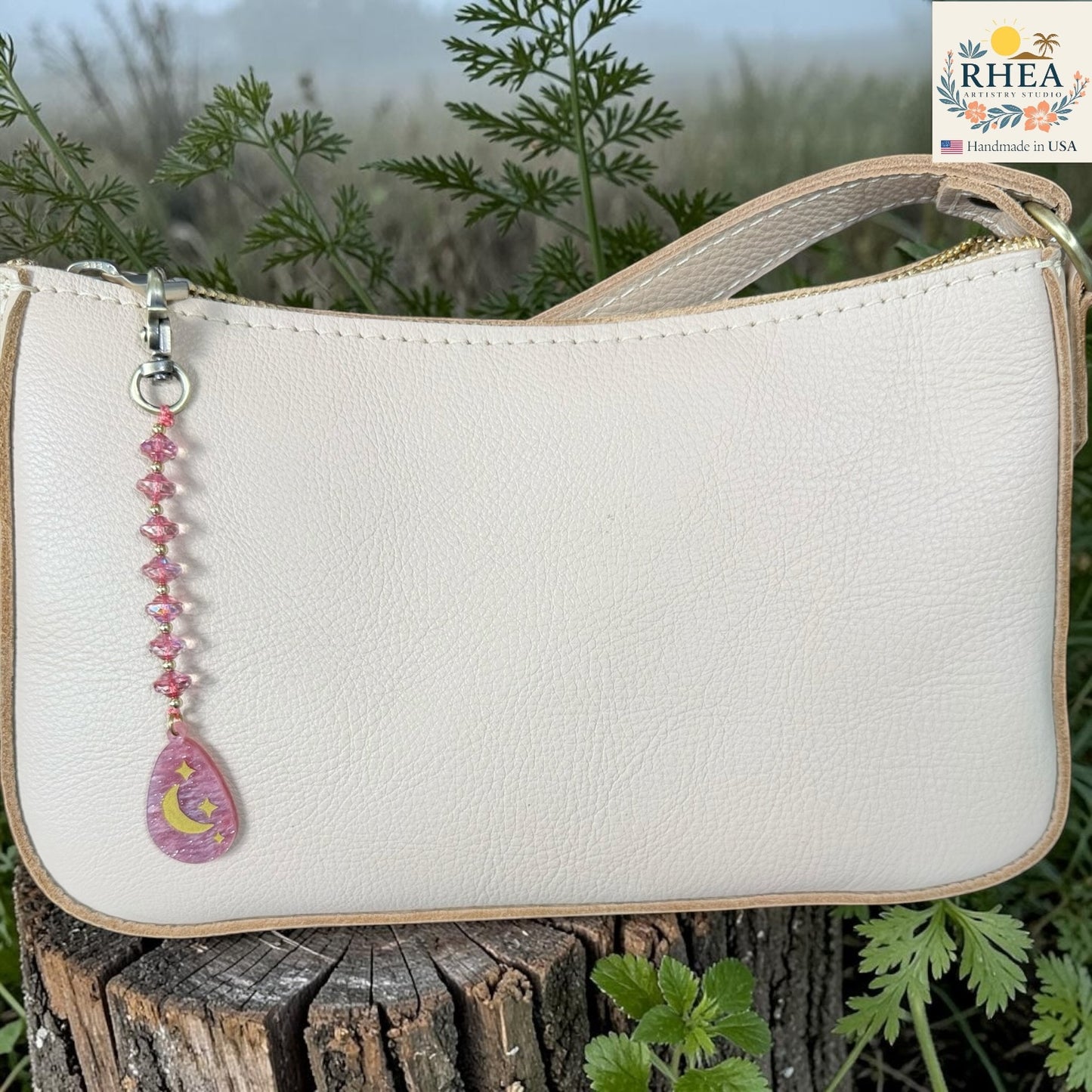 White handbag with a decorative charm on a wooden surface with greenery in the background, featuring the RHEA brand.