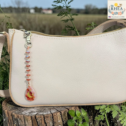 Beige handbag with decorative charm on a wooden stump with plants in the background, featuring the RHEA brand.