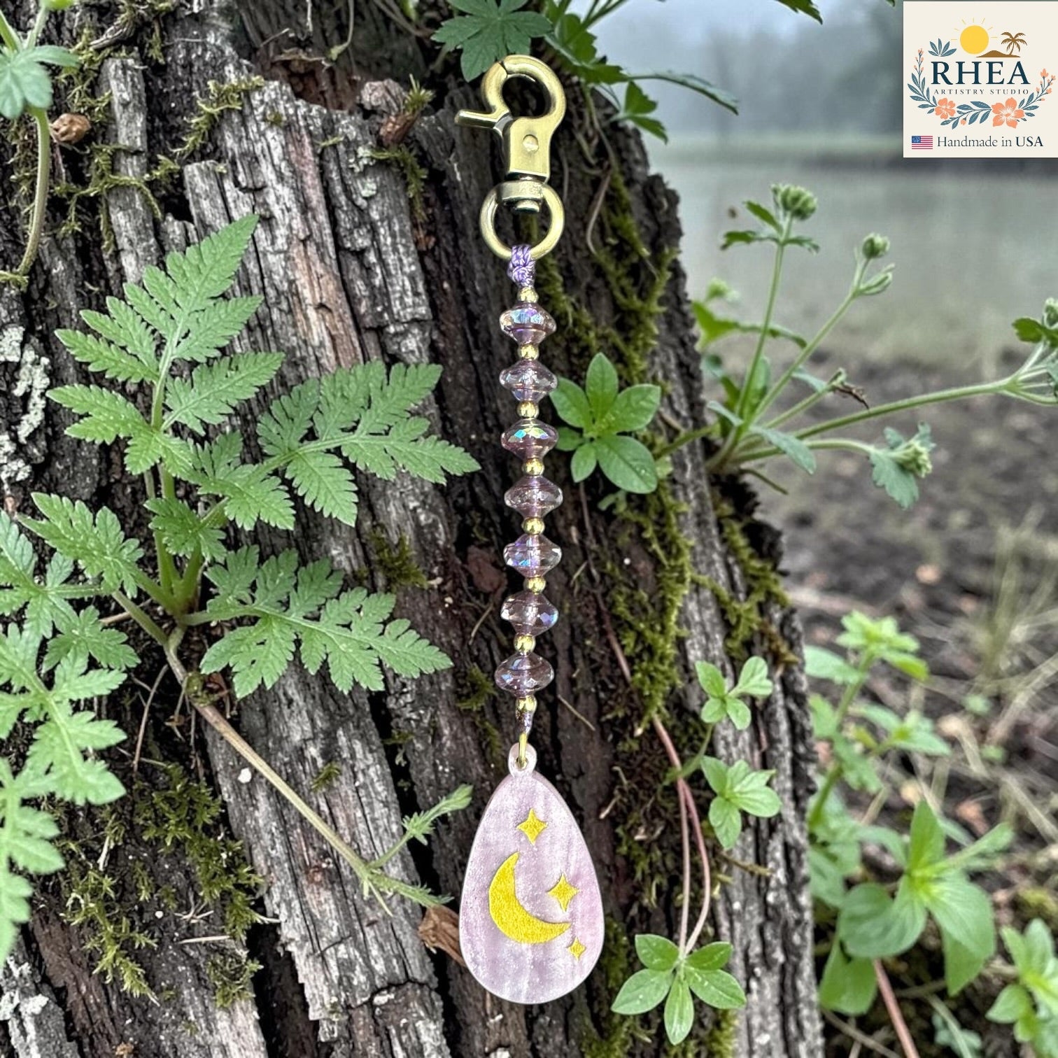 Decorative keychain with moon and star design hanging on a tree branch with plants around.
