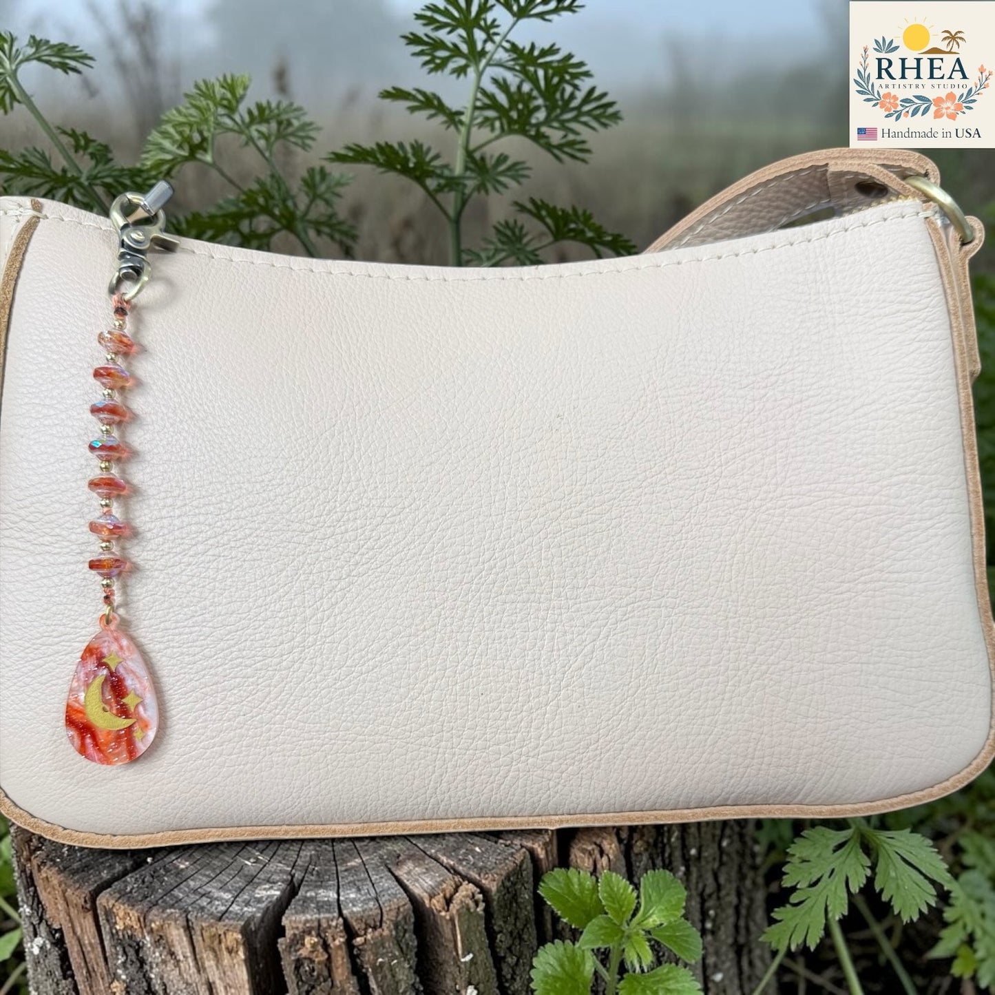 White handbag with a decorative bead chain on a wooden surface with greenery in the background, featuring the RHEA brand.
