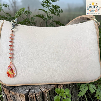 White handbag with a decorative bead chain on a wooden surface with greenery in the background, featuring the RHEA brand.