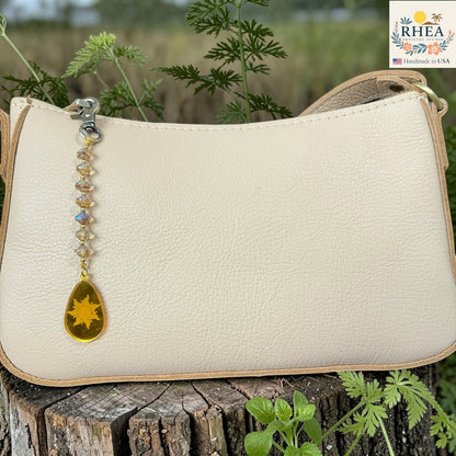 Beige handbag with a decorative charm on a wooden stump with plants in the background, featuring the RHEA brand.