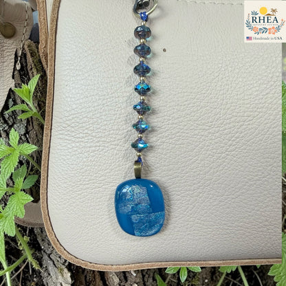 Blue pendant keychain on a textured surface with 'RHEA' branding.
