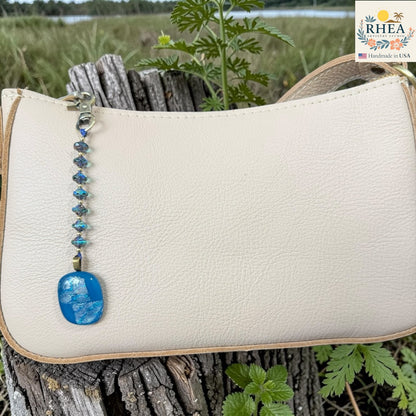 White handbag with a blue pendant on a wooden surface with greenery in the background, featuring the RHEA brand.