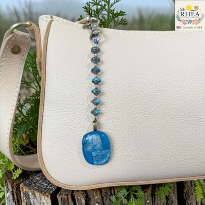 White handbag with a blue beaded keychain on a wooden surface with greenery in the background
