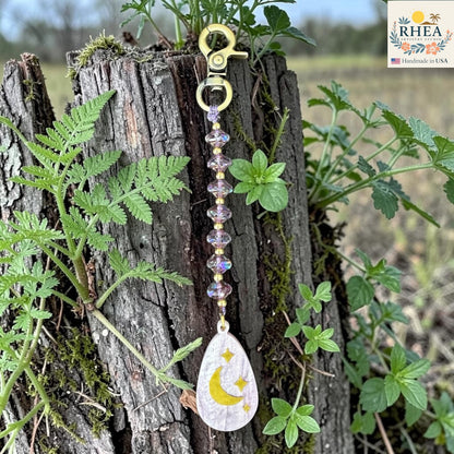 Decorative keychain with moon and star design hanging on a wooden post with plants.