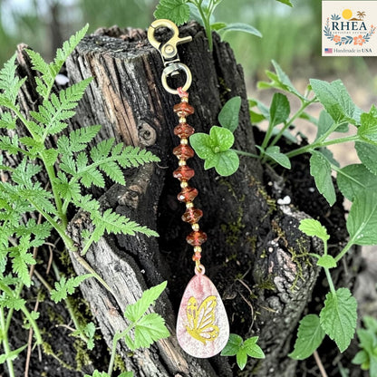 Decorative keychain with butterfly charm on a wooden stump surrounded by greenery, featuring the RHEEA brand.