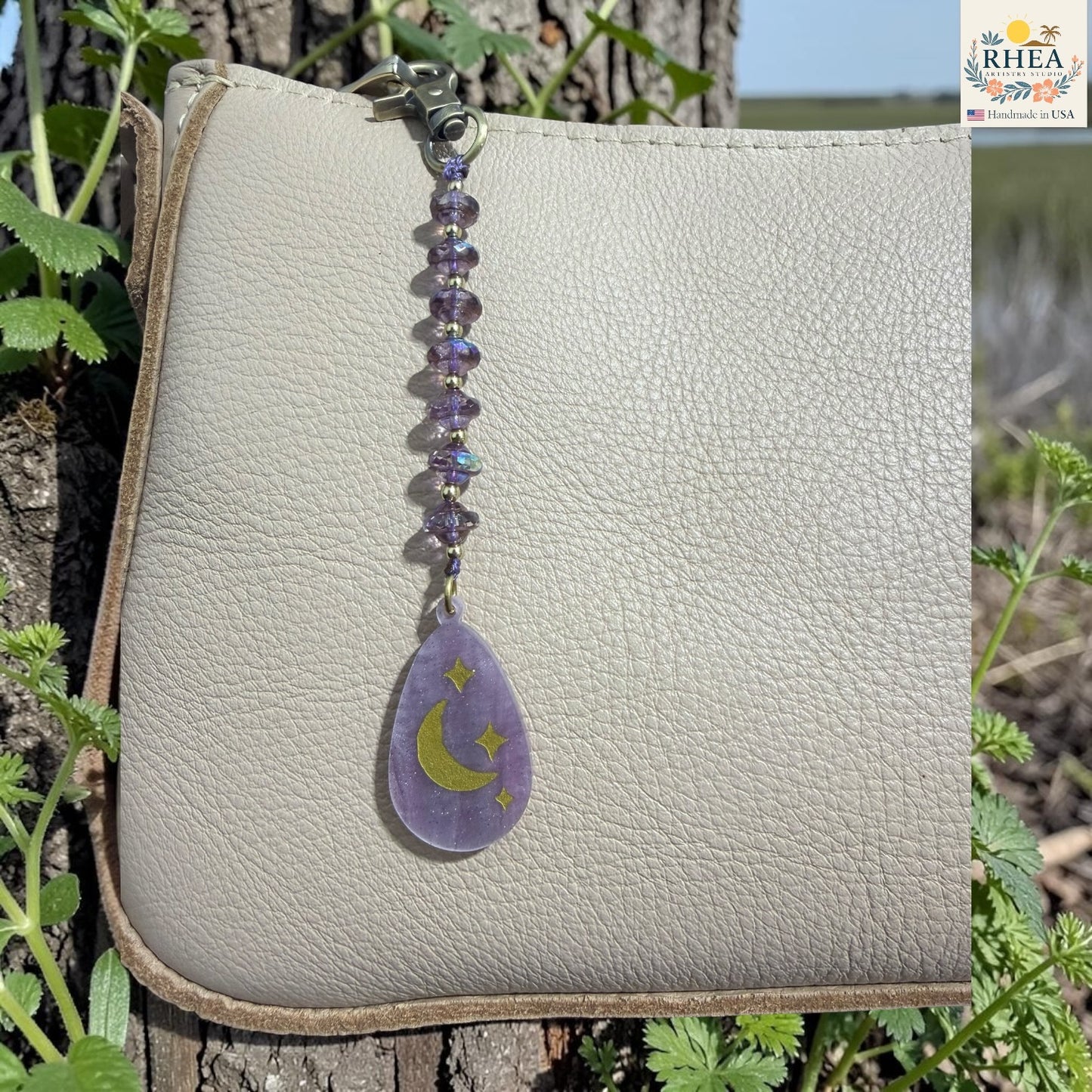 Purple teardrop pendant with moon and star design on a beige leather bag, surrounded by greenery.