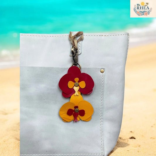 Floral keychain attached to a white bag on a beach background with 'RHEA' branding.