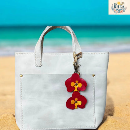 White handbag with a red floral keychain on a sandy beach with blue water in the background, featuring the RHEA brand.