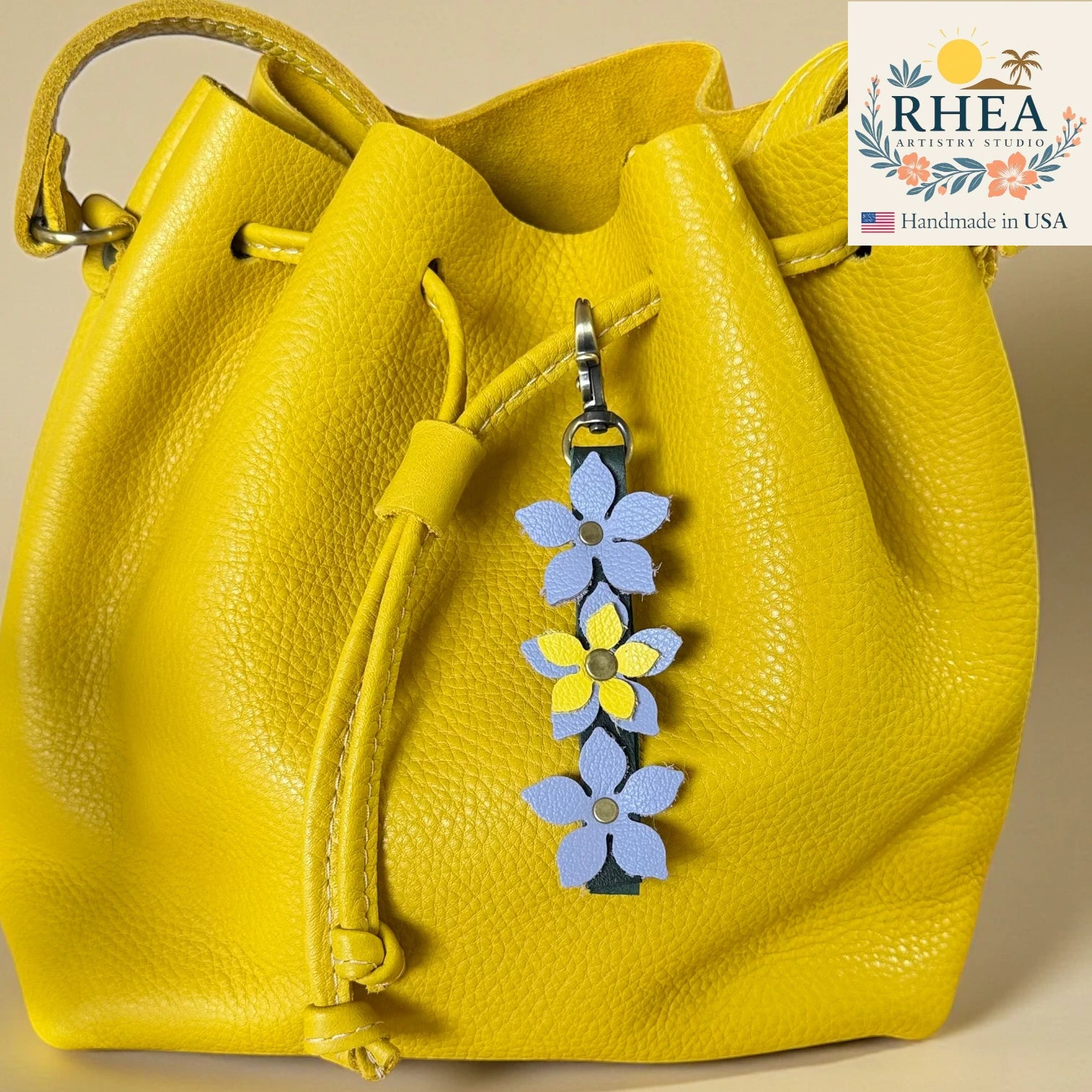 Yellow leather bag with a floral keychain charm on a beige background, featuring the RHEA Artisan Studio logo.