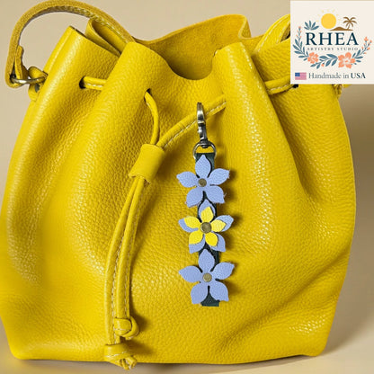 Yellow leather bag with a floral keychain charm on a beige background, featuring the RHEA Artisan Studio logo.