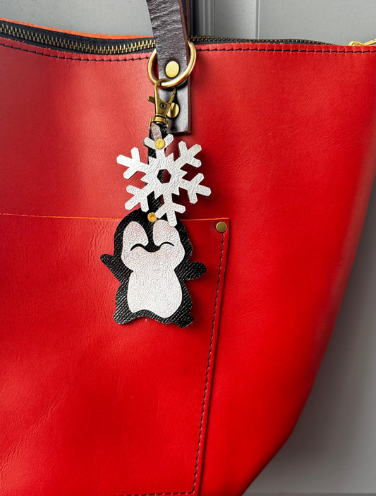 Handcrafted leather charm featuring a laughing penguin with a silver snowflake on top. The charm is designed in a sleek black and silver color combination and includes a lobster clasp for easy attachment to purses or use as a keychain.