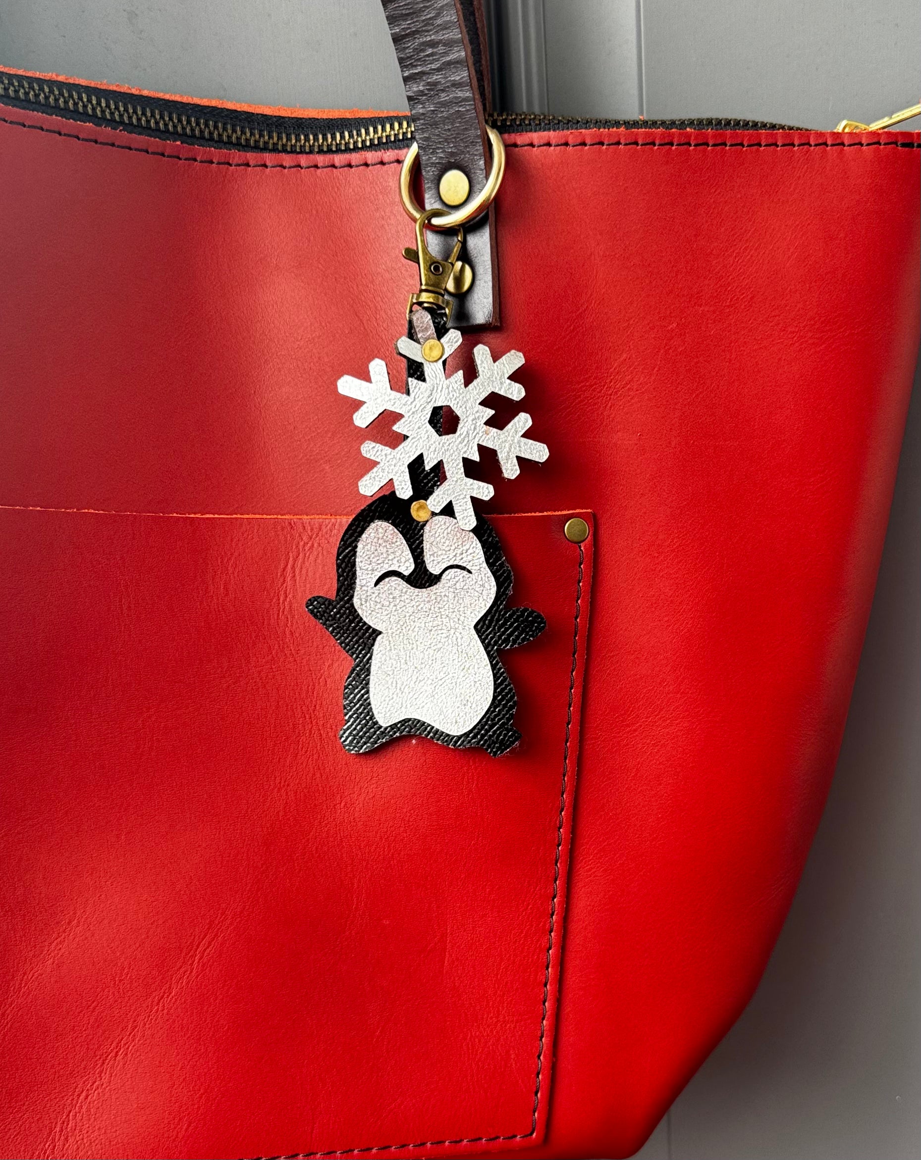 Handcrafted leather charm featuring a laughing penguin with a silver snowflake on top. The charm is designed in a sleek black and silver color combination and includes a lobster clasp for easy attachment to purses or use as a keychain.