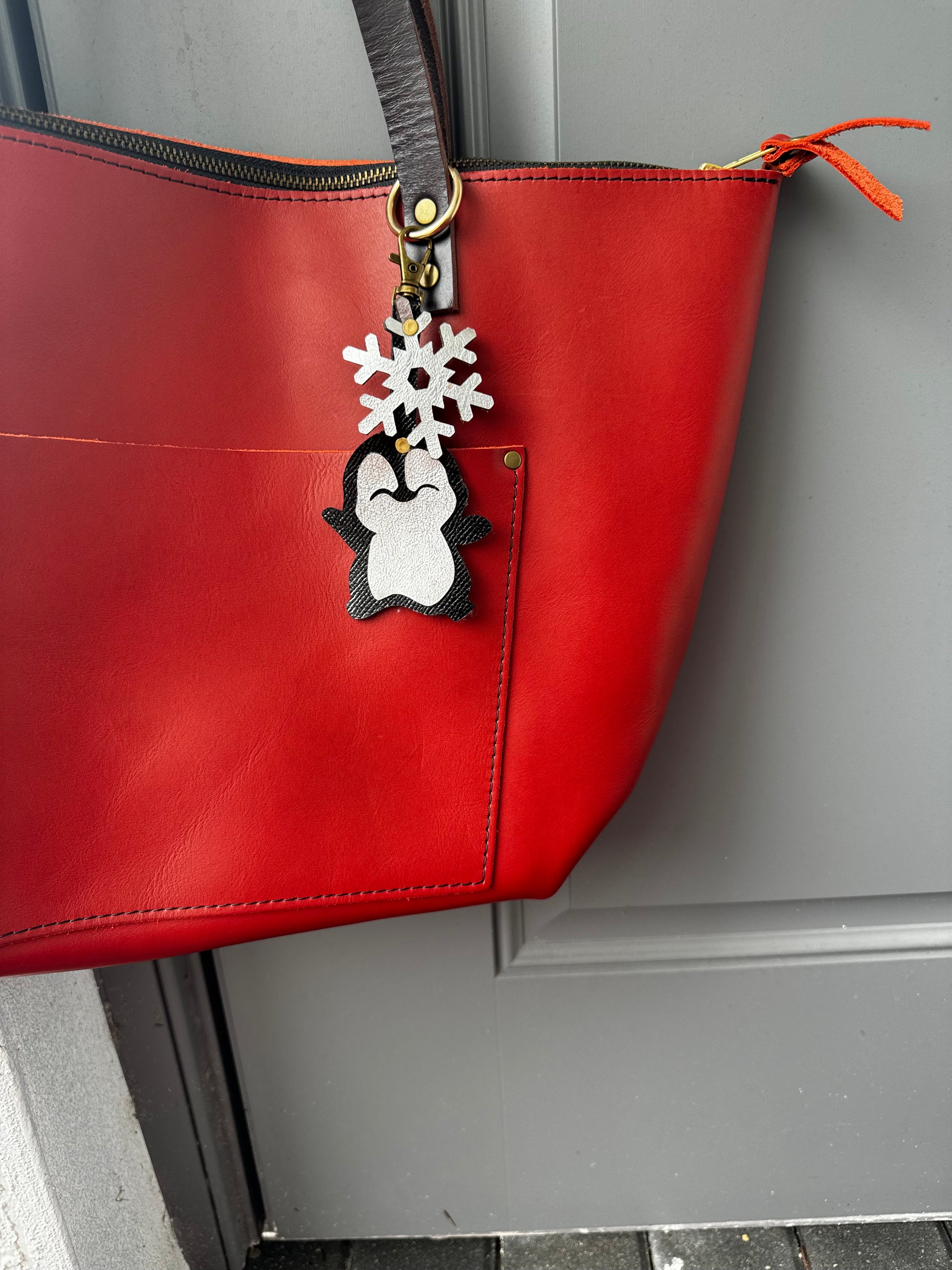 Handcrafted leather charm featuring a laughing penguin with a silver snowflake on top. The charm is designed in a sleek black and silver color combination and includes a lobster clasp for easy attachment to purses or use as a keychain.