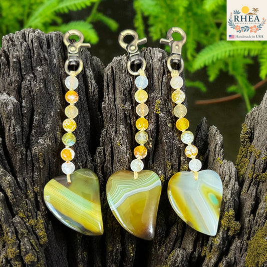 Three heart-shaped stone pendants with beaded extensions on a wooden surface, branded 'RHEEA'.