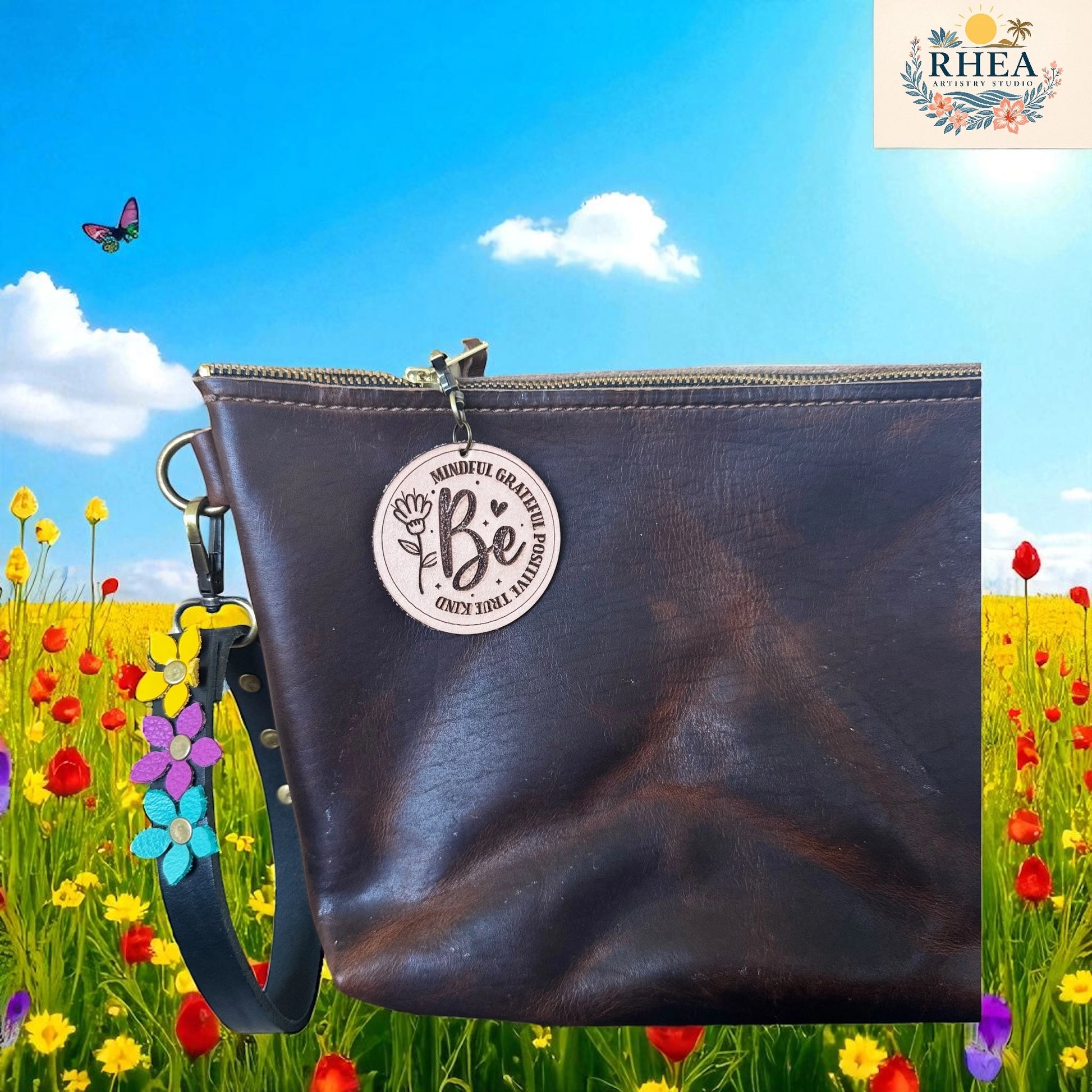 Brown leather clutch bag with 'Be' charm in a field of flowers and butterflies, featuring the RHEEA brand.