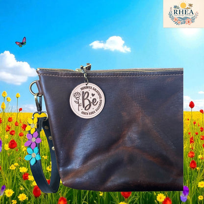 Brown leather clutch bag with 'Be' charm in a field of flowers and butterflies, featuring the RHEEA brand.