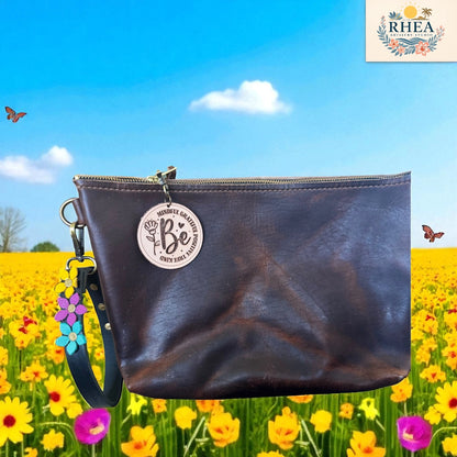 Brown leather pouch with 'Be' charm on a background of a blue sky and yellow flowers.