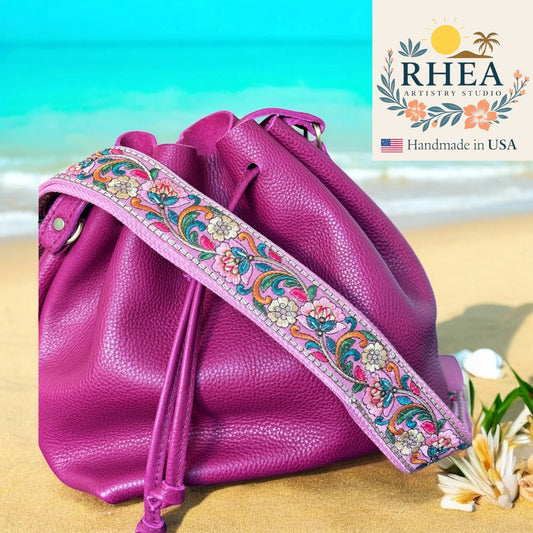 Pink leather bag with a colorful strap on a beach, featuring RHEEA Artistry Studio logo.
