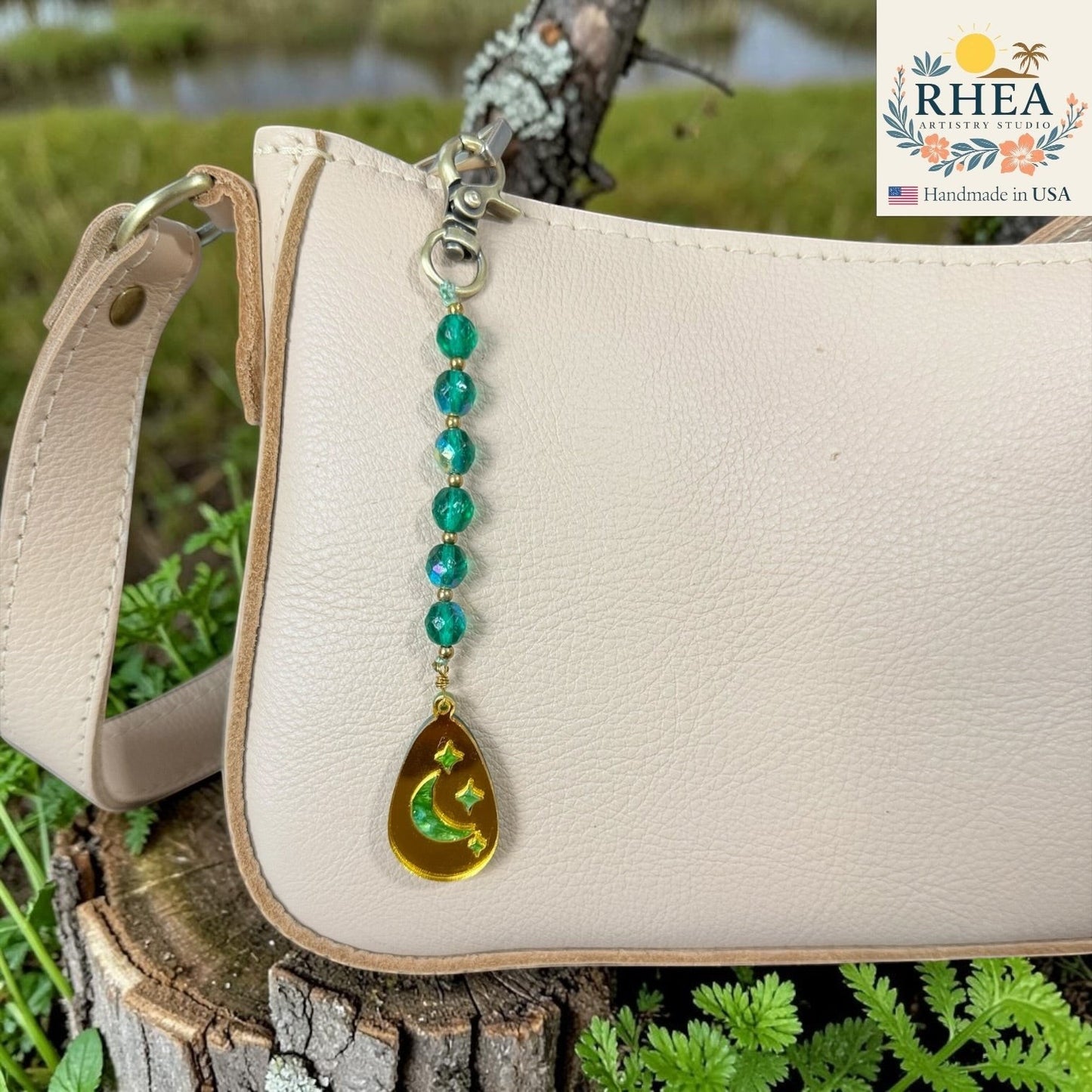 Beaded keychain with a gold teardrop charm on a beige bag, with RHEA branding in the corner.
