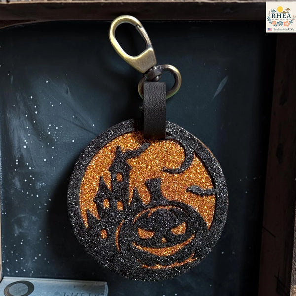 A round Halloween-themed keychain or bag charm with a black glitter border, orange glitter center, black silhouette of a haunted house and jack-o'-lantern, attached to a black strap and gold-tone clasp, shown against a dark backdrop.