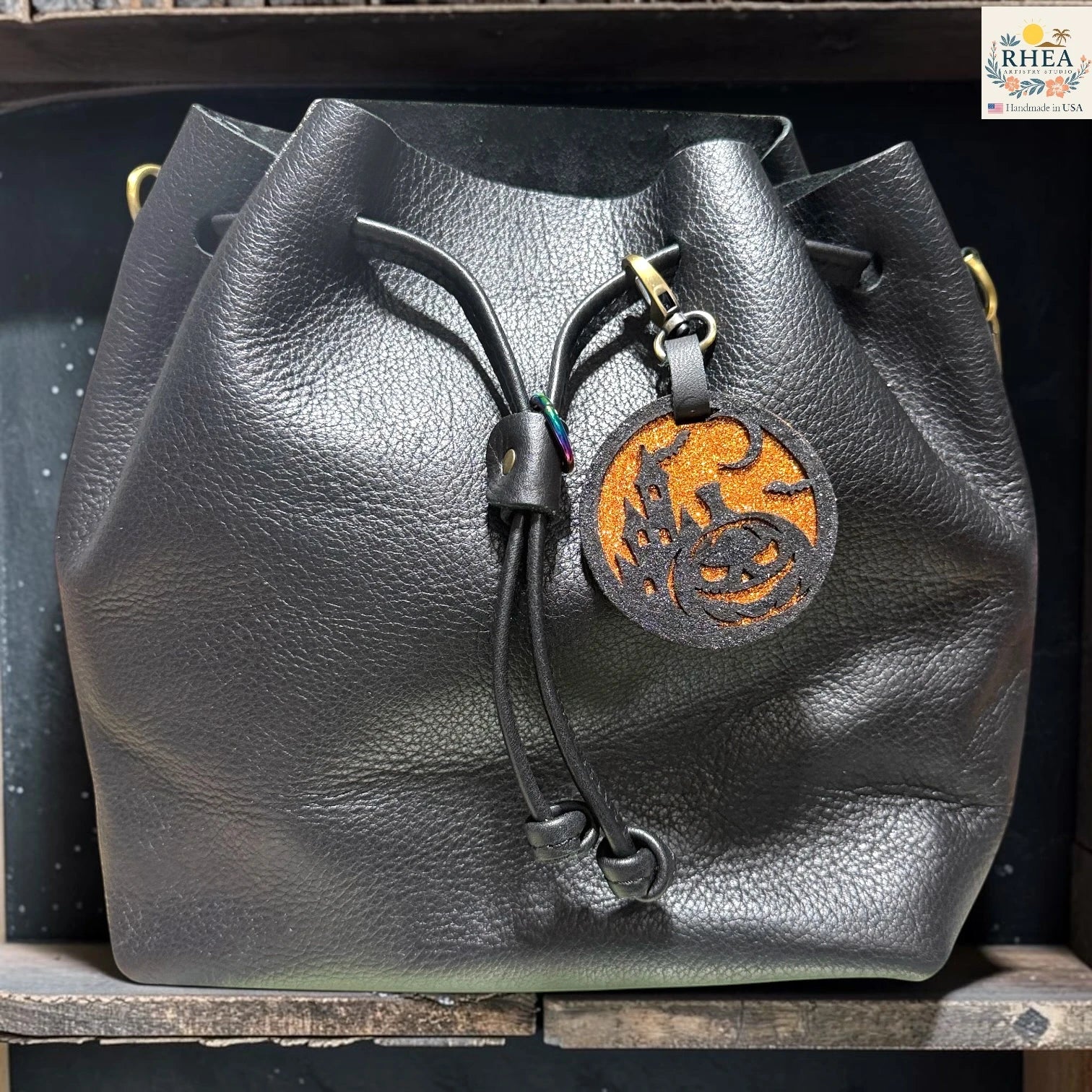 Black leather bucket bag with a drawstring closure and a circular leather charm featuring a stylized orange and black design, displayed on a wooden shelf.