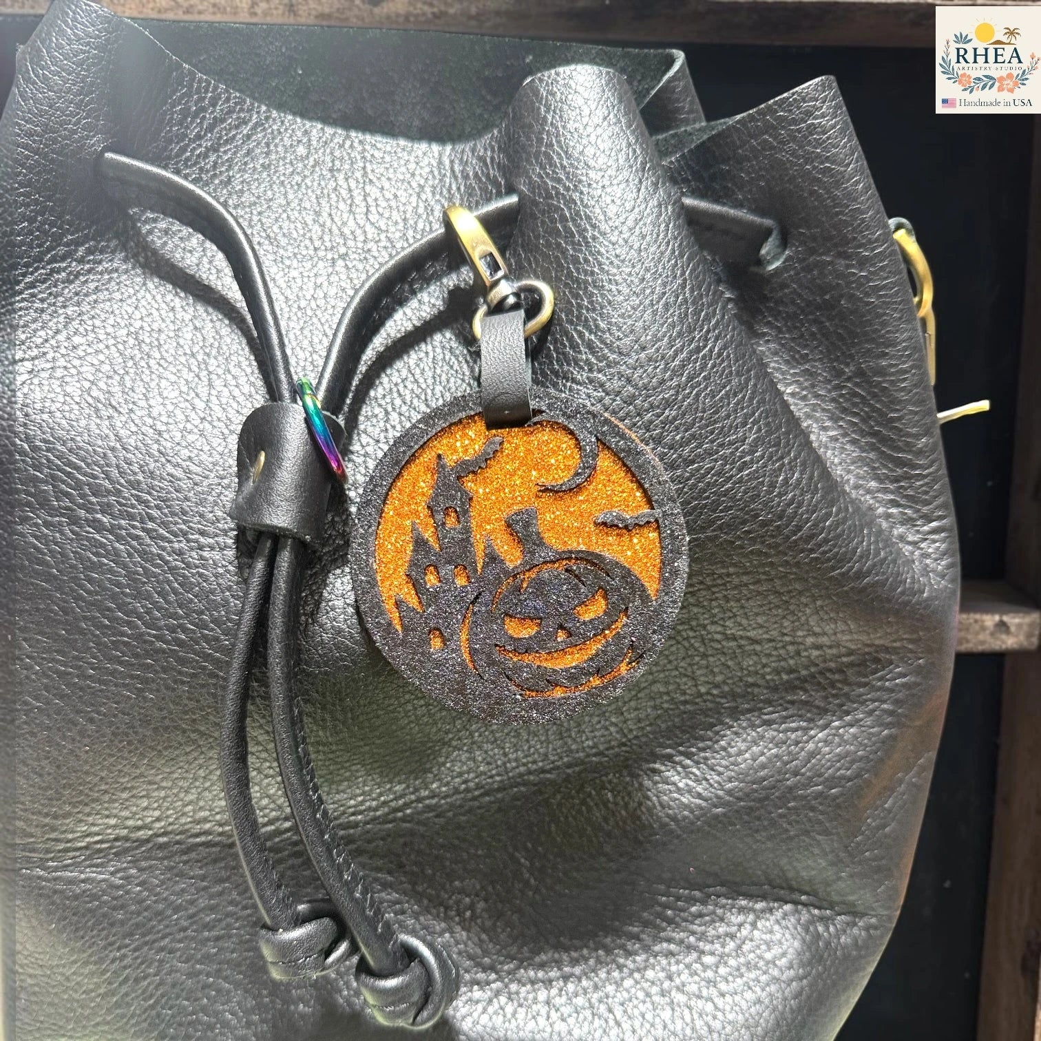 Black pebbled leather bucket bag with a drawstring closure and a circular orange and black Halloween-themed hanging charm featuring a jack-o'-lantern and crescent moon.