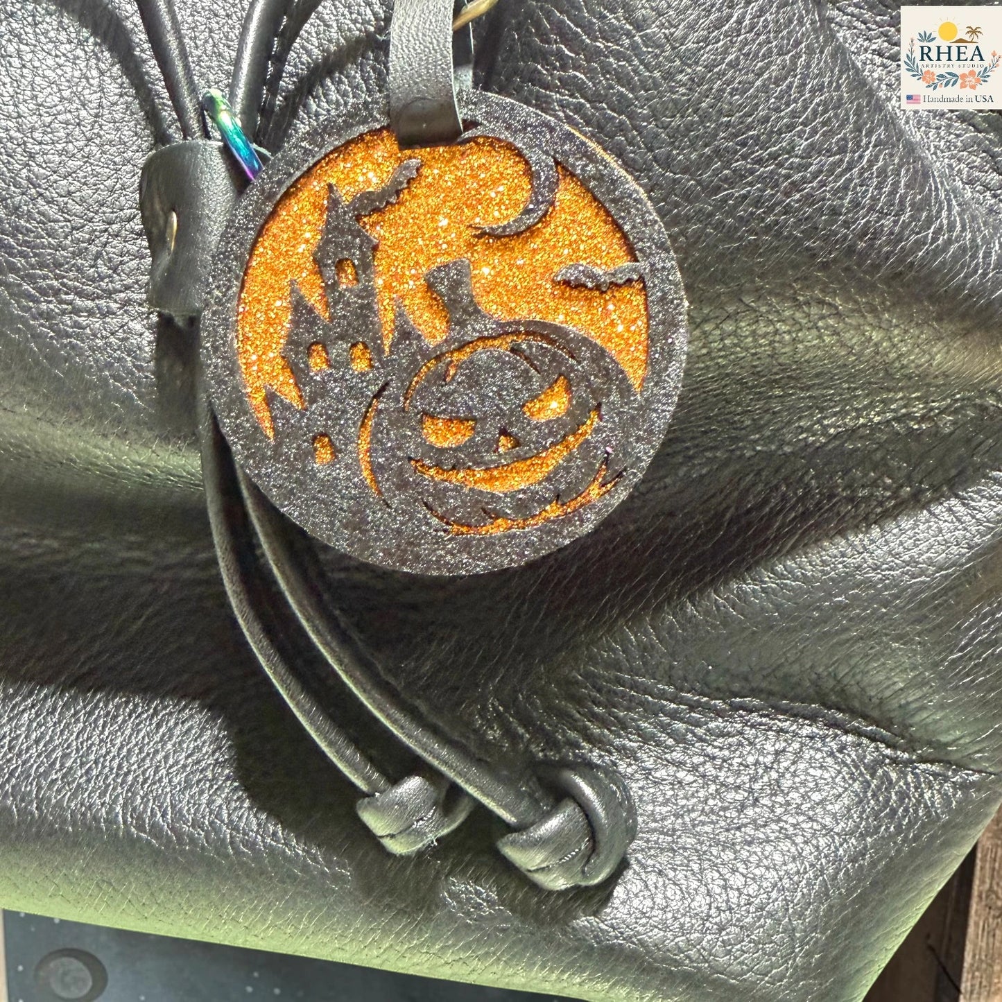 A Halloween-themed leather keychain or bag charm with a glittery orange circular disc featuring a black jack-o'-lantern and haunted house design, attached to black leather cords, shown resting on a black textured leather surface.
