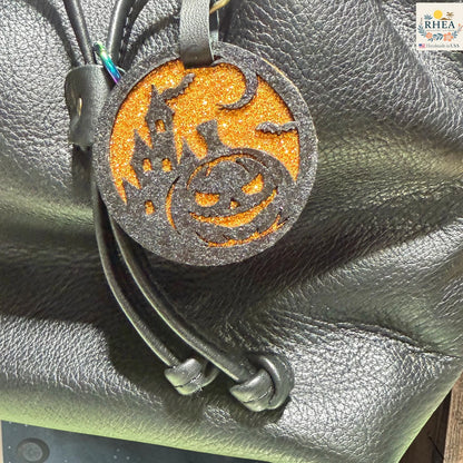 A Halloween-themed leather keychain or bag charm with a glittery orange circular disc featuring a black jack-o'-lantern and haunted house design, attached to black leather cords, shown resting on a black textured leather surface.