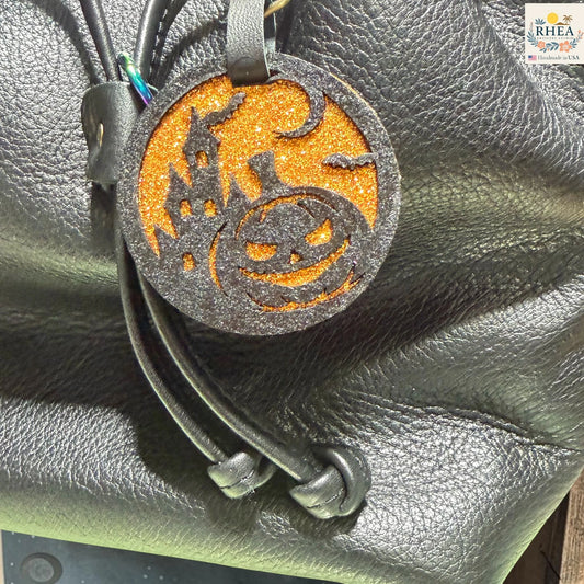 A Halloween-themed leather keychain or bag charm with a glittery orange circular disc featuring a black jack-o'-lantern and haunted house design, attached to black leather cords, shown resting on a black textured leather surface.