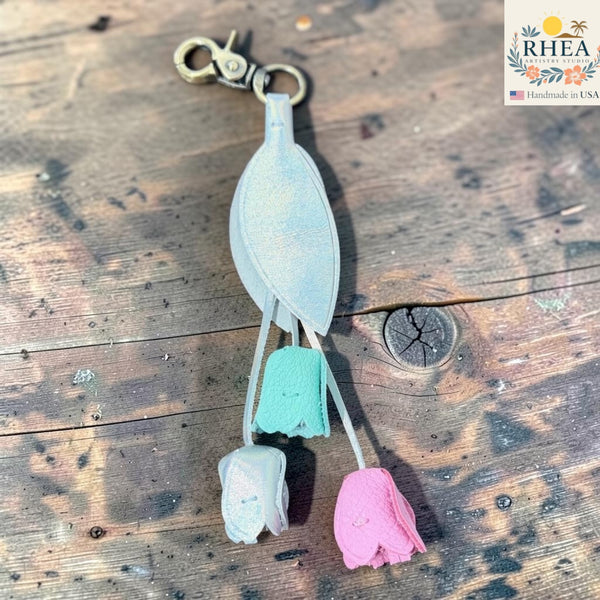 A handmade keychain featuring a silver-toned leaf charm with three dangling tulip charms in pastel pink, mint green, and white, resting on a rustic wooden surface. The brand logo 'RHEA Handmade in USA' is visible in the top right corner.