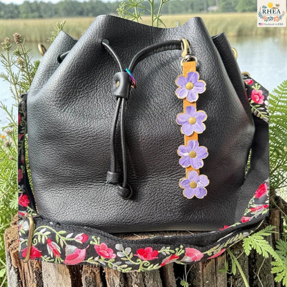 Rhea Artistry Studio Hand-Painted Leather Flower Bag Charm in Periwinkle Meadow with periwinkle blue and soft lavender painted leather flowers, brass rivets, honey leather strap, and antique brass clasp.