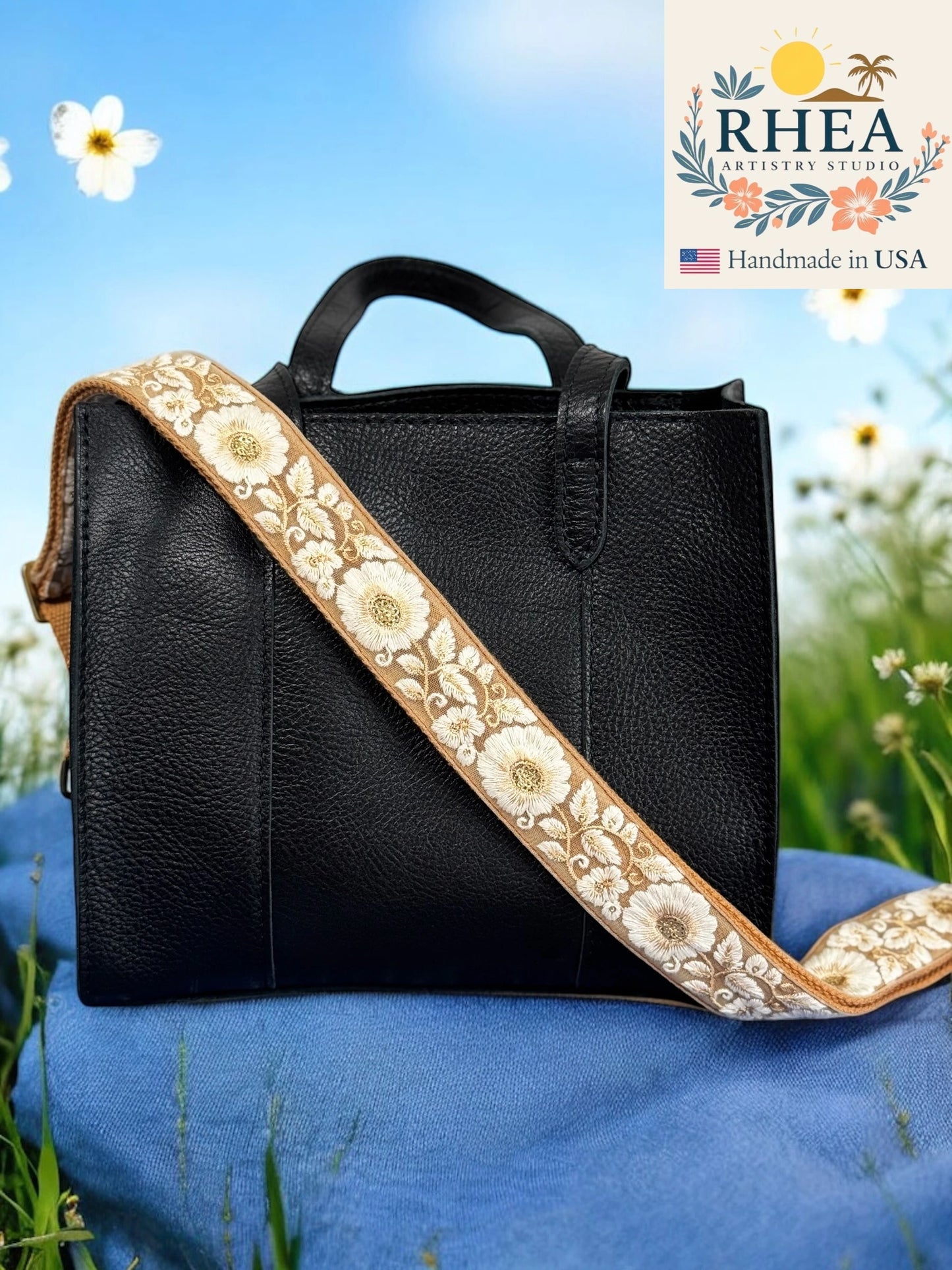 Black handbag with floral purse strap on a blue cushion against a natural background, featuring the RHEA Artistry Studio logo.