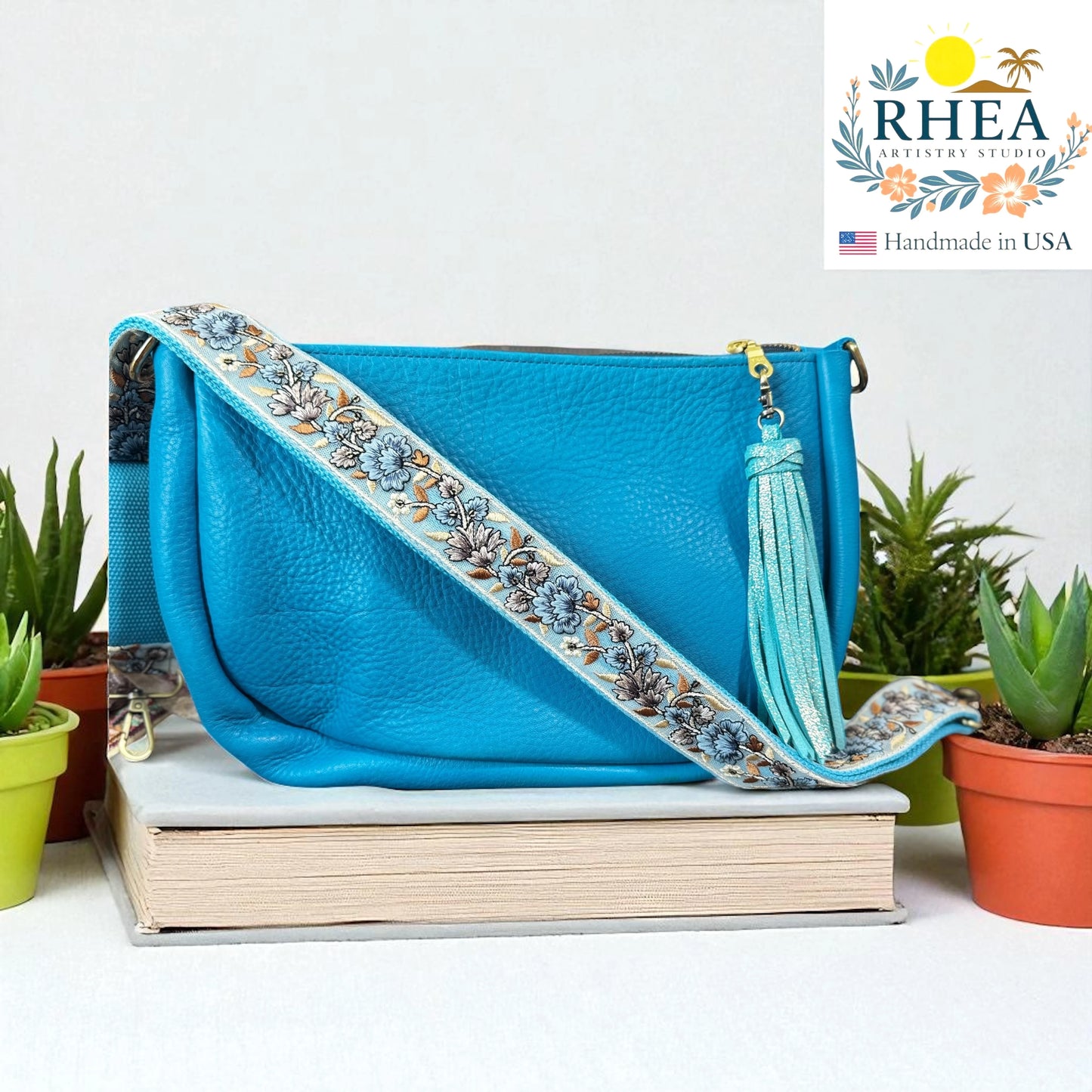 Blue handbag with a blue floral strap on a stack of books, with RHEA branding in the corner.