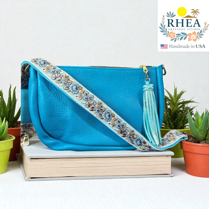Blue handbag with a blue floral strap on a stack of books, with RHEA branding in the corner.