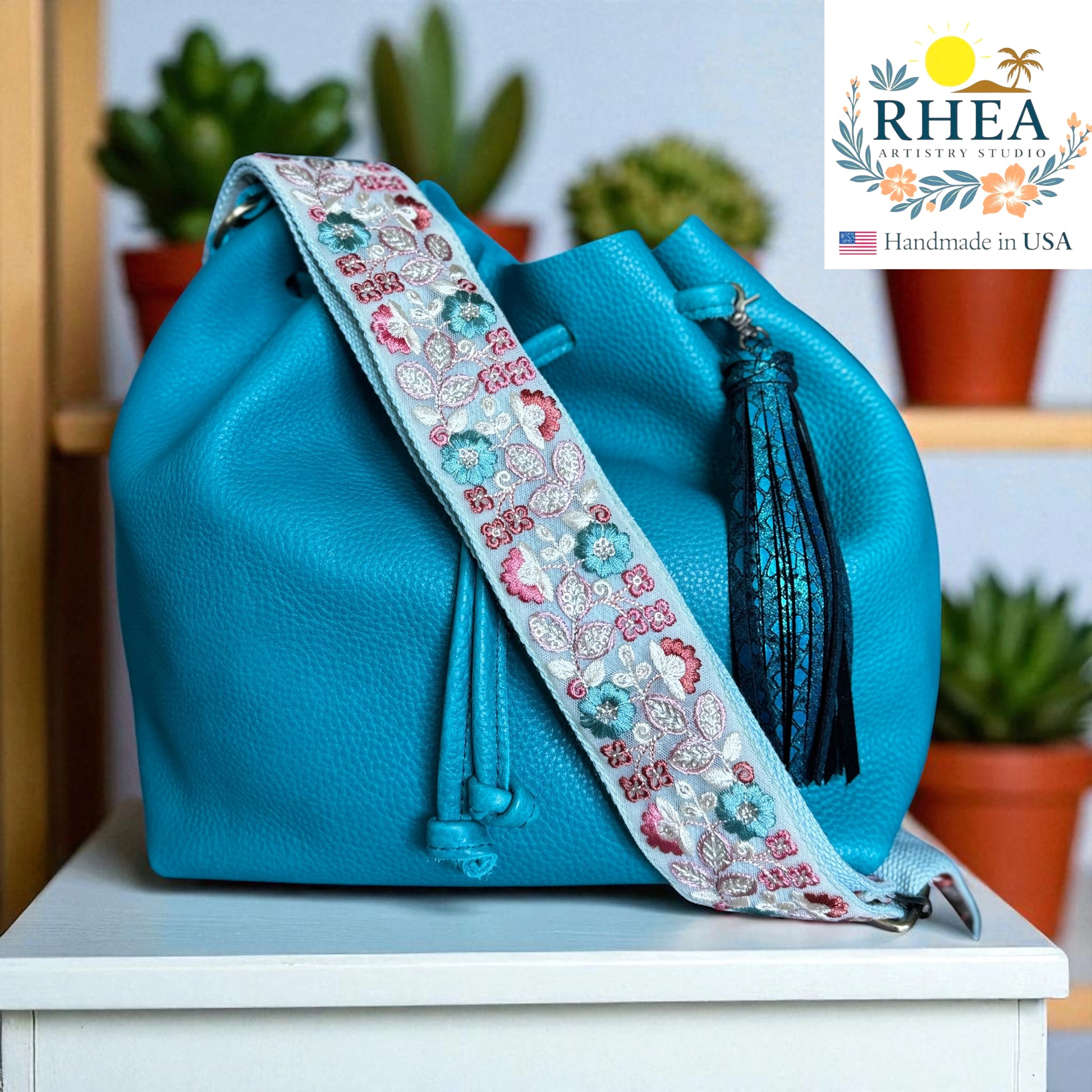 Blue handbag with a decorative strap on a white surface, with 'RHEA' branding in the corner.