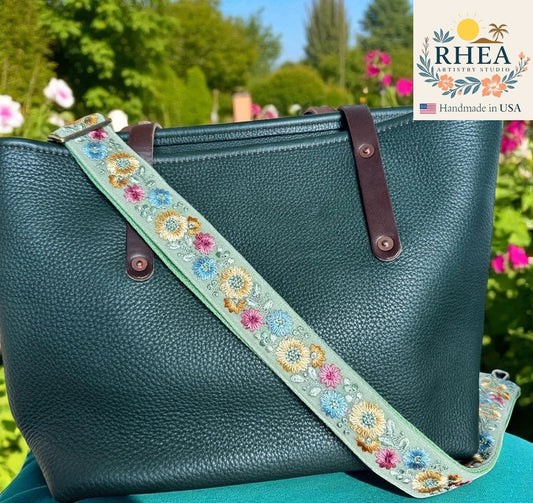 Teal handbag with a floral embroidered strap against a garden background, featuring the RHEA Artisan Studio logo.