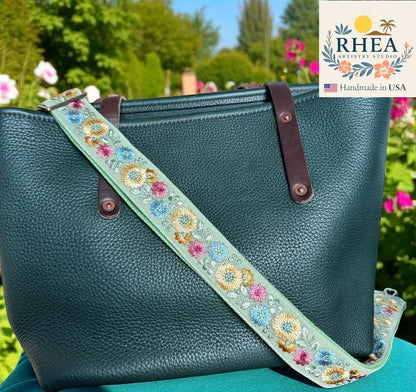 Teal handbag with floral strap against a garden background, featuring the RHEA brand.