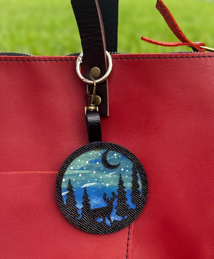 A handpainted leather charm with a starry night landscape, a deer, and a forest design, attached to a red leather bag.