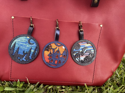 A handpainted leather charm with a starry night landscape, a deer, and a forest design, attached to a red leather bag.