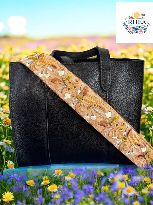 Embroidered bag strap featuring golden wheat-like motifs, curling vines, and cream florals on a warm tan background, styled on a black leather bag.