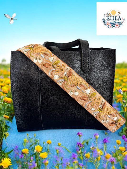 Embroidered bag strap featuring golden wheat-like motifs, curling vines, and cream florals on a warm tan background, styled on a black leather bag.