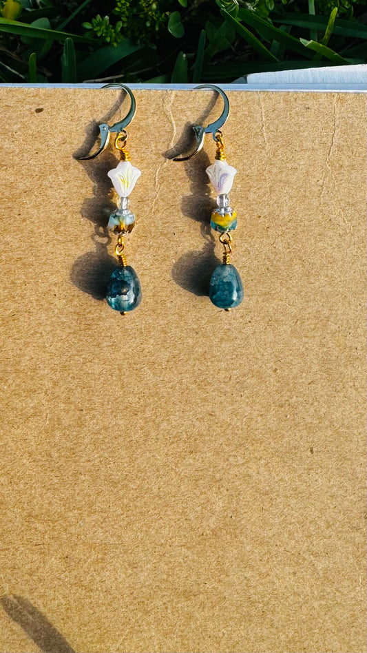 A pair of gold-colored three petal cathedral drop earrings with white and blue czech glass accents, displayed against a beige background.